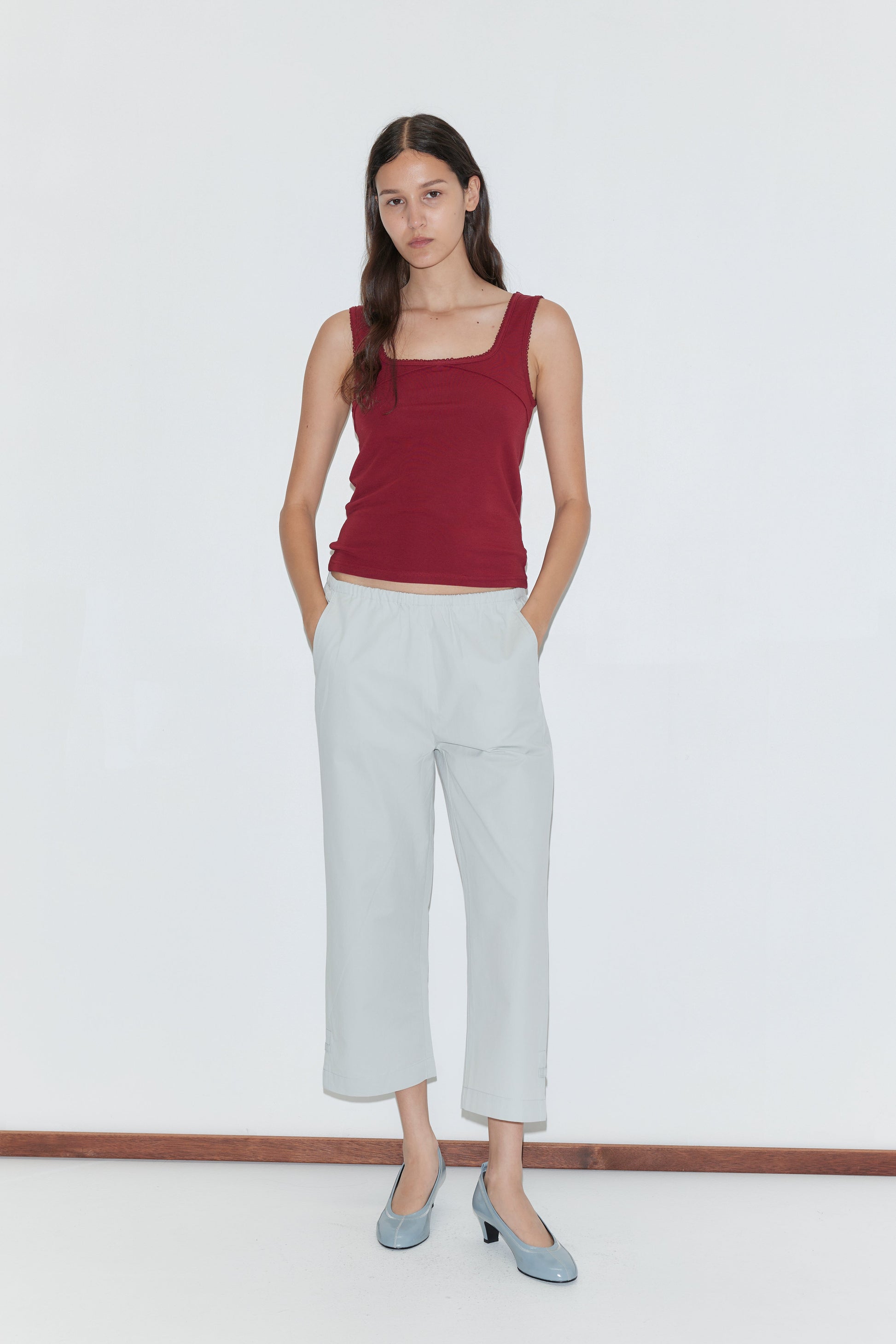 A woman stands against a white background, wearing a sleeveless maroon top and Baby Blue Capri Pant with an elastic waistband, hands in her pockets and a neutral expression.