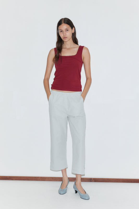 A woman stands against a white background, wearing a sleeveless maroon top and Baby Blue Capri Pant with an elastic waistband, hands in her pockets and a neutral expression.