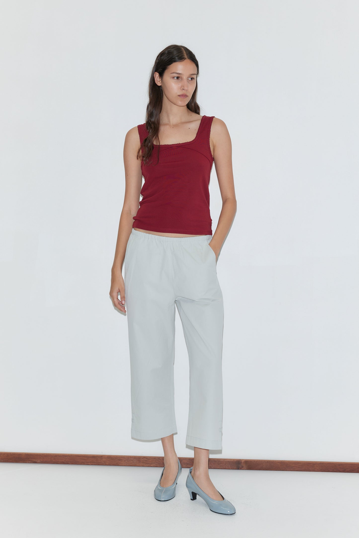 A woman with long dark hair wears a sleeveless maroon top, Capri Pant - Baby Blue with an elasticated waistband, and light blue heels. She stands against a plain white background with one hand in her pocket, looking to the side.