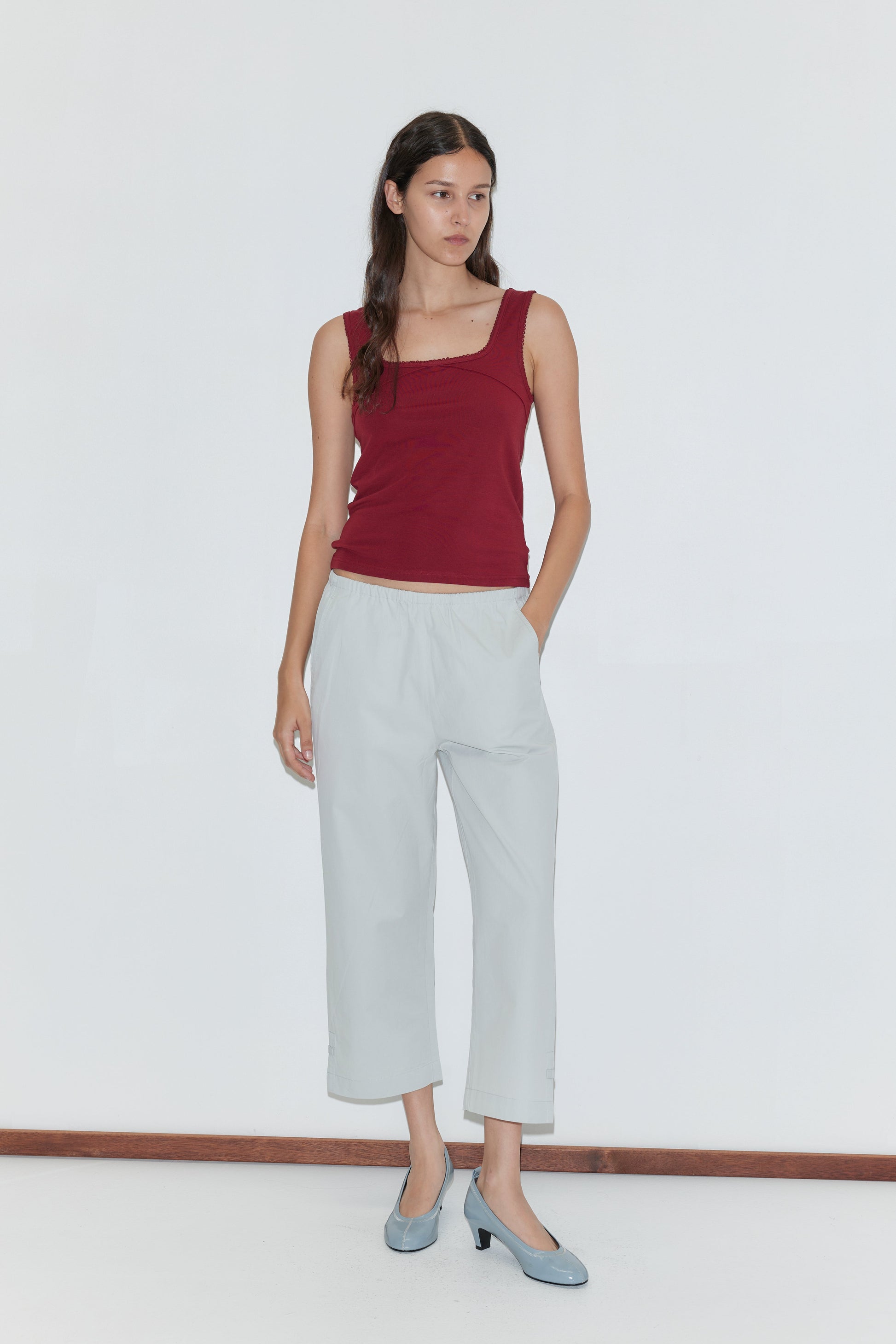 A woman with long dark hair wears a sleeveless maroon top, Capri Pant - Baby Blue with an elasticated waistband, and light blue heels. She stands against a plain white background with one hand in her pocket, looking to the side.