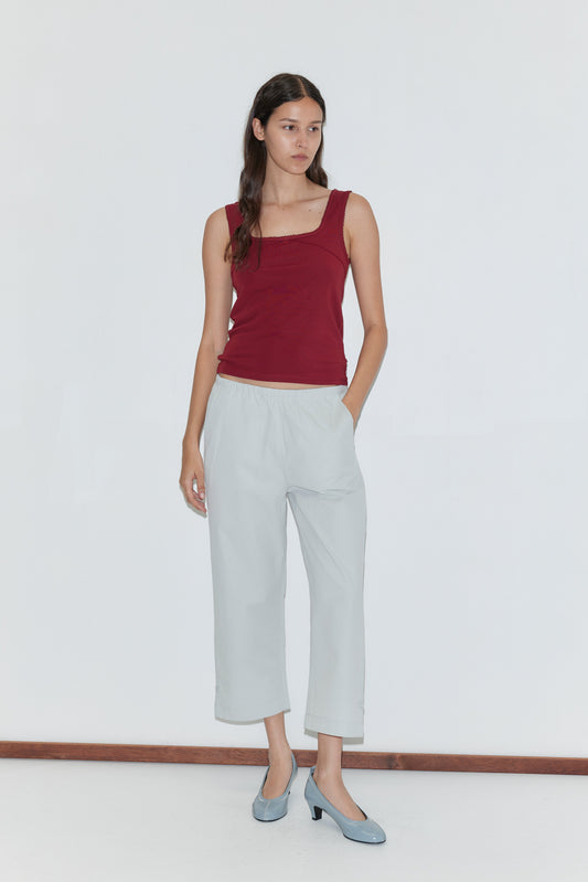A woman with long dark hair wears a sleeveless maroon top, Capri Pant - Baby Blue with an elasticated waistband, and light blue heels. She stands against a plain white background with one hand in her pocket, looking to the side.