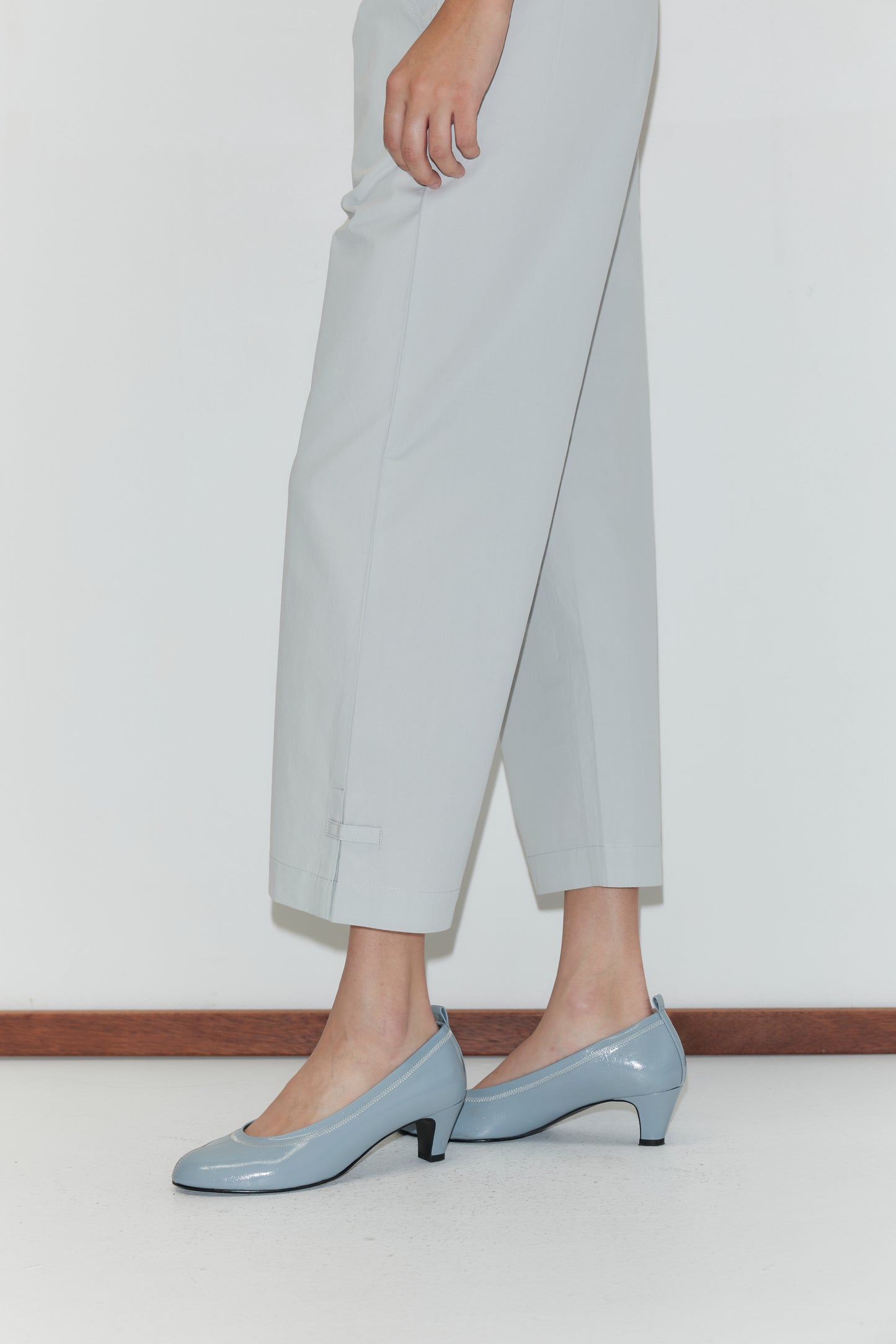 A person stands indoors wearing the Capri Pant - Baby Blue, which features a relaxed fit and elasticated waistband, paired with light blue low-heeled shoes. The photo captures their lower half against a white wall and floor with wooden trim.