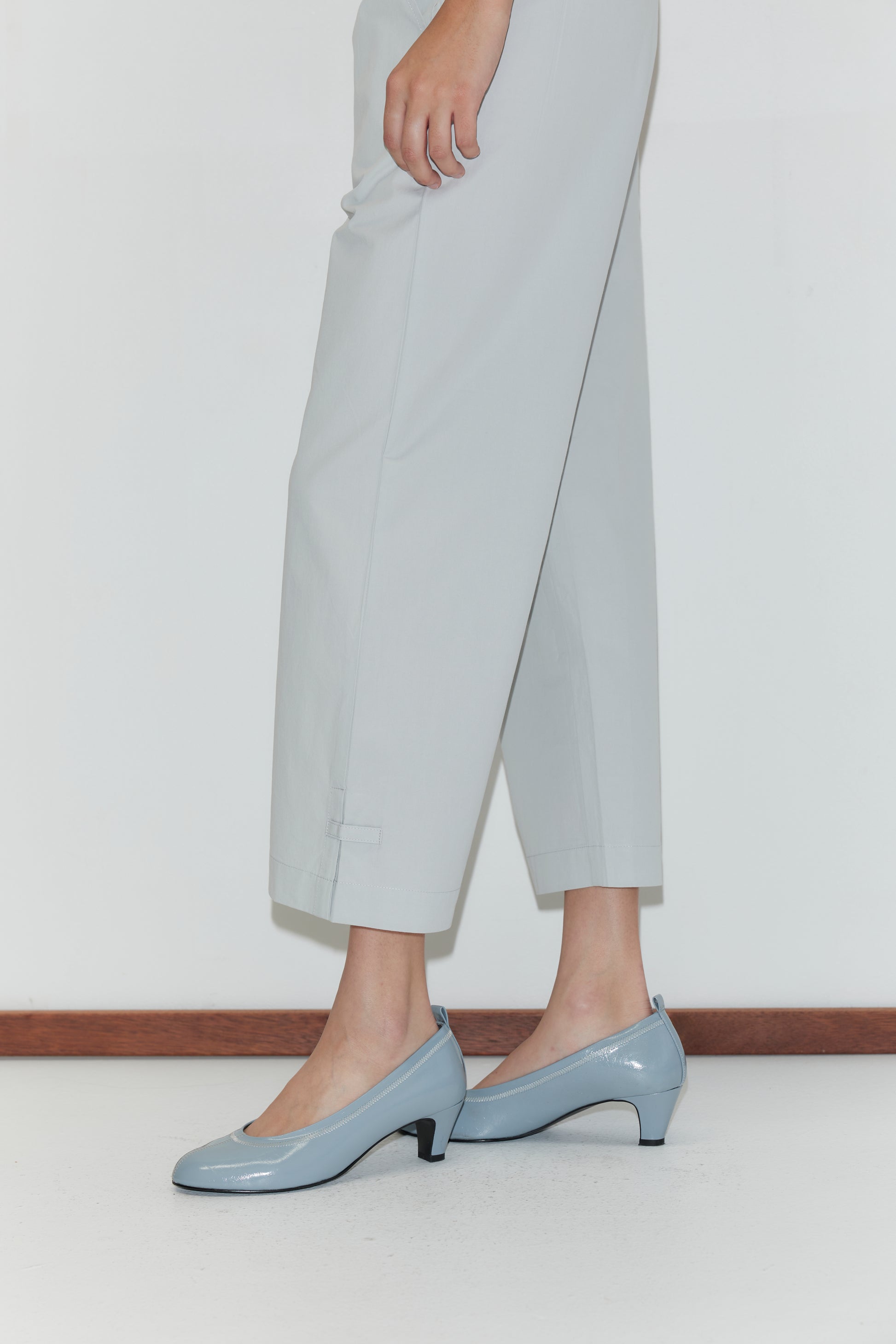 A person stands indoors wearing the Capri Pant - Baby Blue, which features a relaxed fit and elasticated waistband, paired with light blue low-heeled shoes. The photo captures their lower half against a white wall and floor with wooden trim.