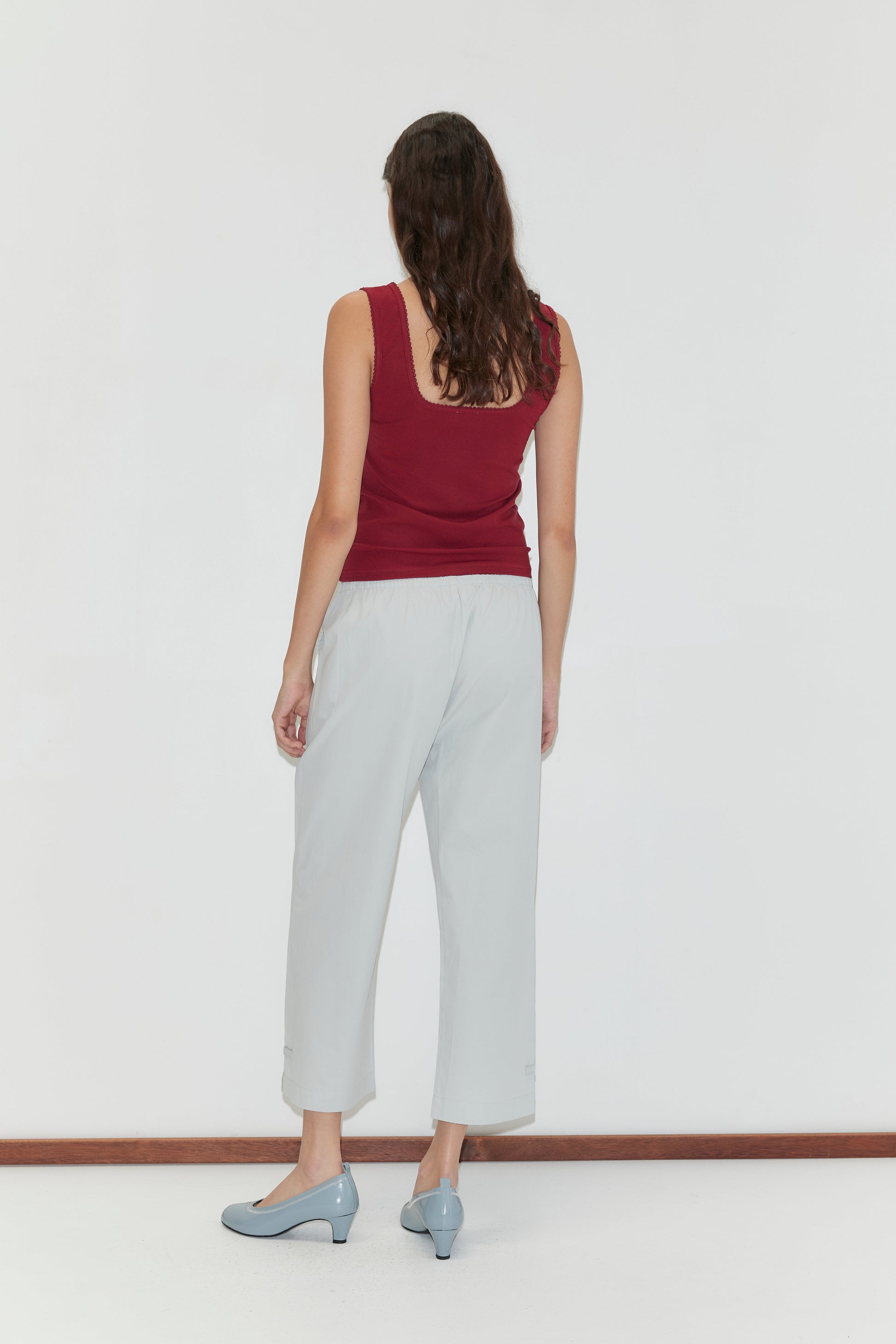 A woman with long brown hair, wearing a sleeveless maroon top and Capri Pant - Baby Blue, stands facing a white wall with her back to the camera, her light blue low-heeled shoes visible.