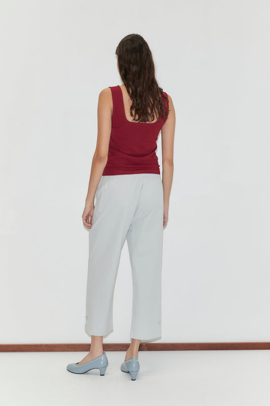 A woman with long brown hair, wearing a sleeveless maroon top and Capri Pant - Baby Blue, stands facing a white wall with her back to the camera, her light blue low-heeled shoes visible.