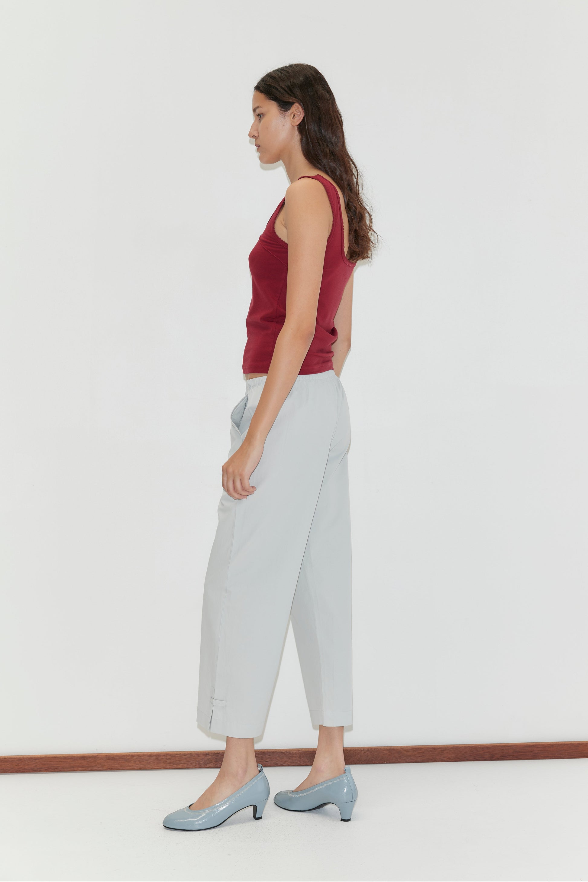 A woman with long, wavy brown hair stands in profile against a white background, wearing a sleeveless maroon top, Capri Pant - Baby Blue with an elasticated waistband, and light blue heels.