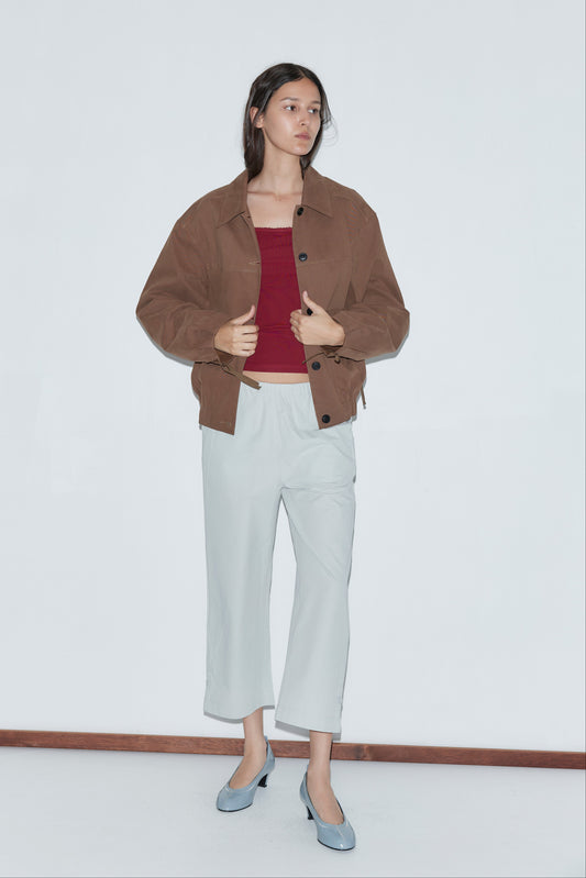 A woman with long dark hair and a neutral expression stands against a white wall wearing the Button Up Jacket - Brown by Deiji Studios, paired with a red top, light gray cropped pants, and light blue heels. She holds one side of her jacket.