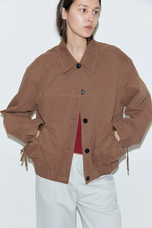 A person wears the Button Up Jacket - Brown over a red top and light gray pants, standing against a plain white background with hands in the jacket pockets.