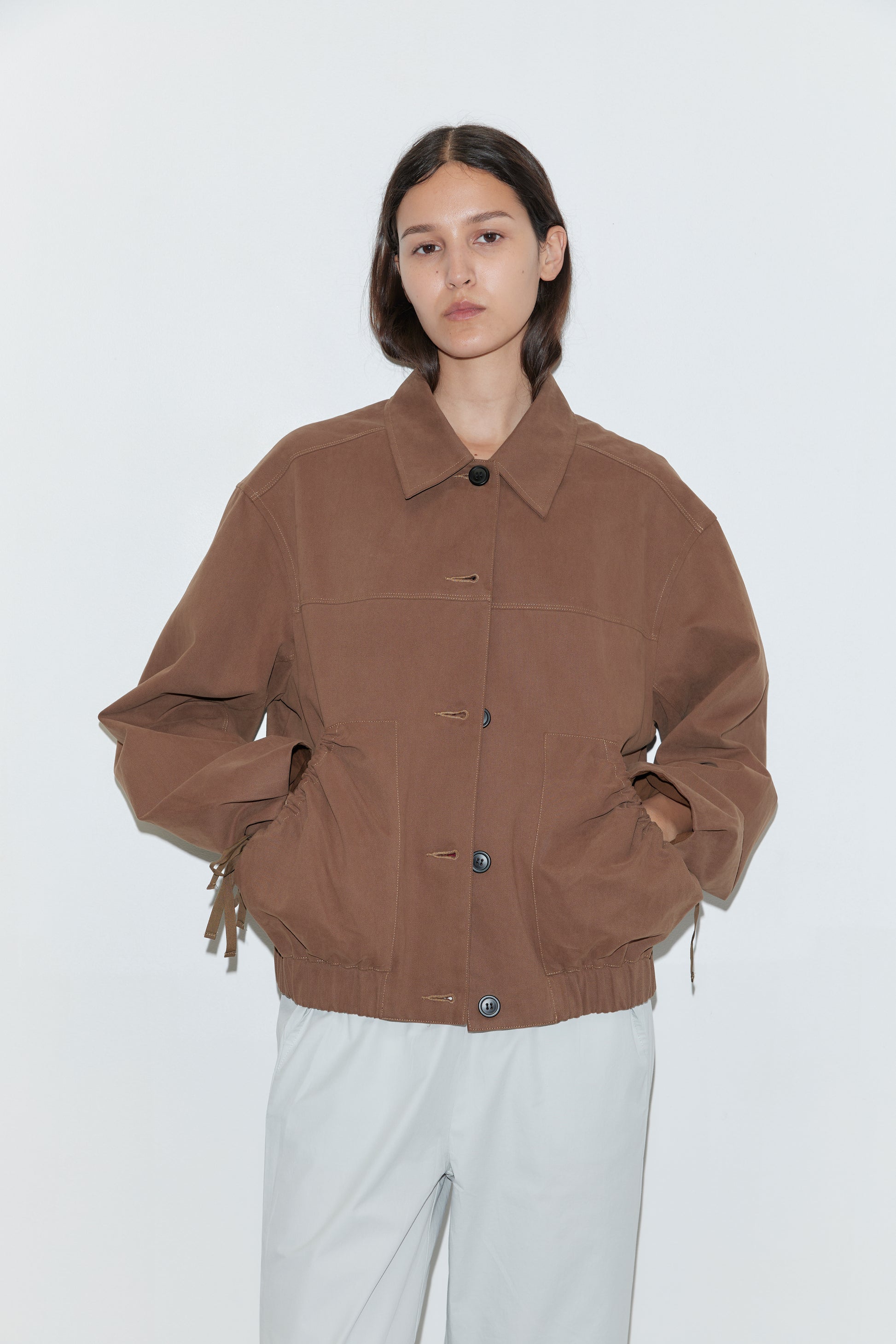 A person with long, dark hair stands against a white background, wearing a loose-fitting Button Up Jacket - Brown with large pockets and light gray pants. Their hands are tucked into the jacket's pockets.