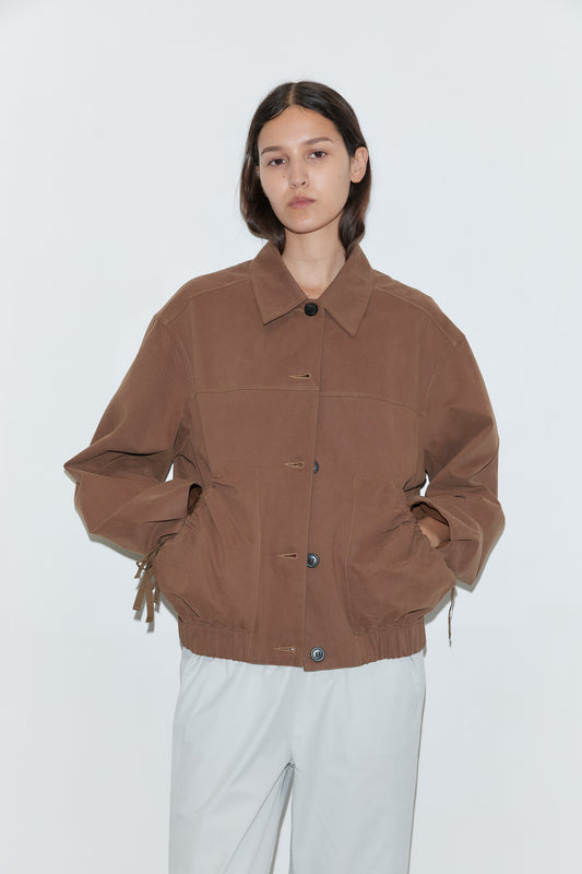 A person with long, dark hair stands against a white background, wearing a loose-fitting Button Up Jacket - Brown with large pockets and light gray pants. Their hands are tucked into the jacket's pockets.