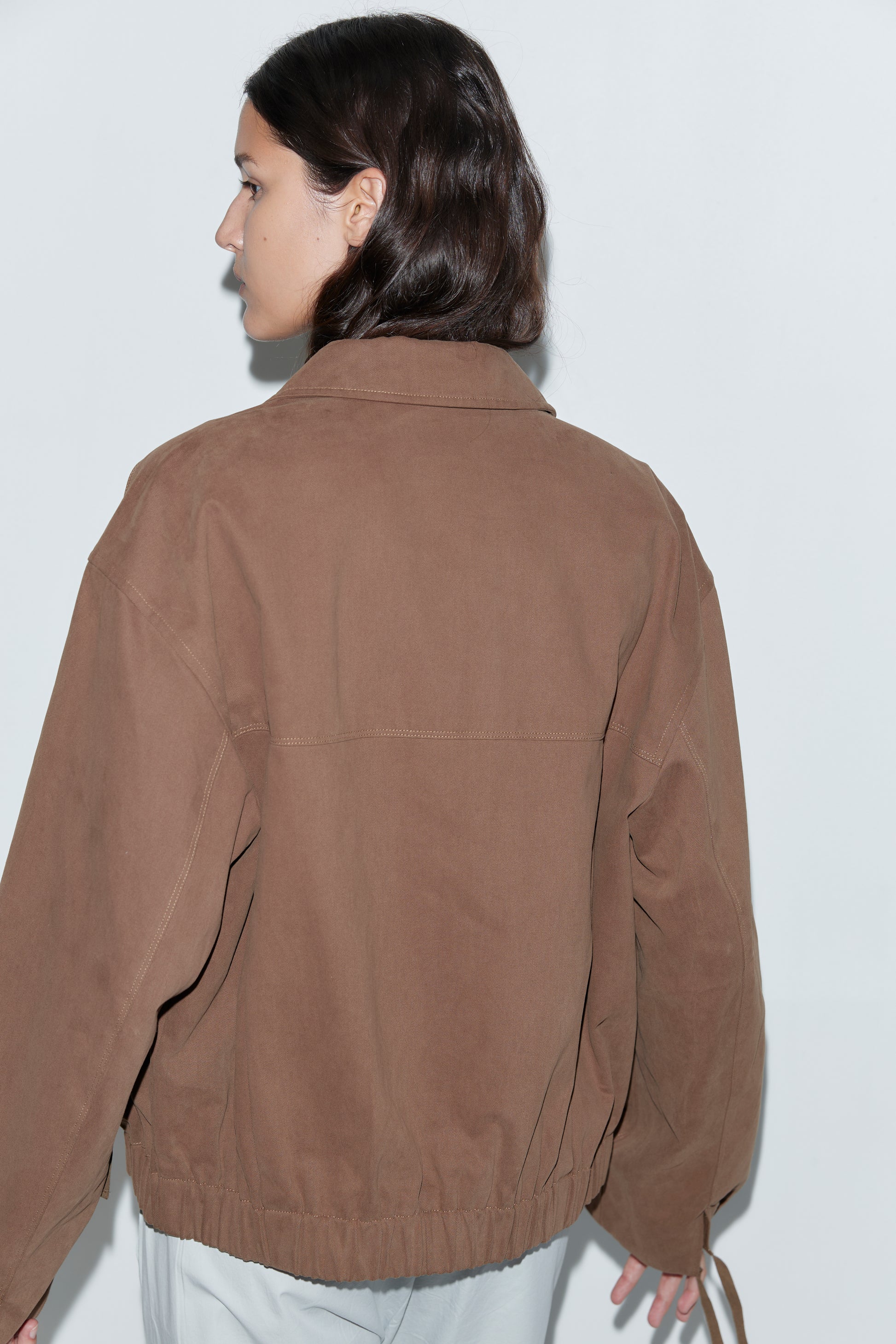 A person with medium-length brown hair is seen from behind wearing the Button Up Jacket - Brown and light pants, standing against a plain white background.