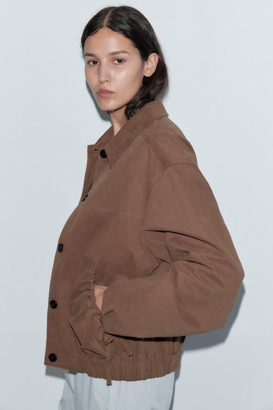 A person with long dark hair wears the Button Up Jacket - Brown from Deiji Studios and light pants, standing calmly against a plain white background and looking at the camera.