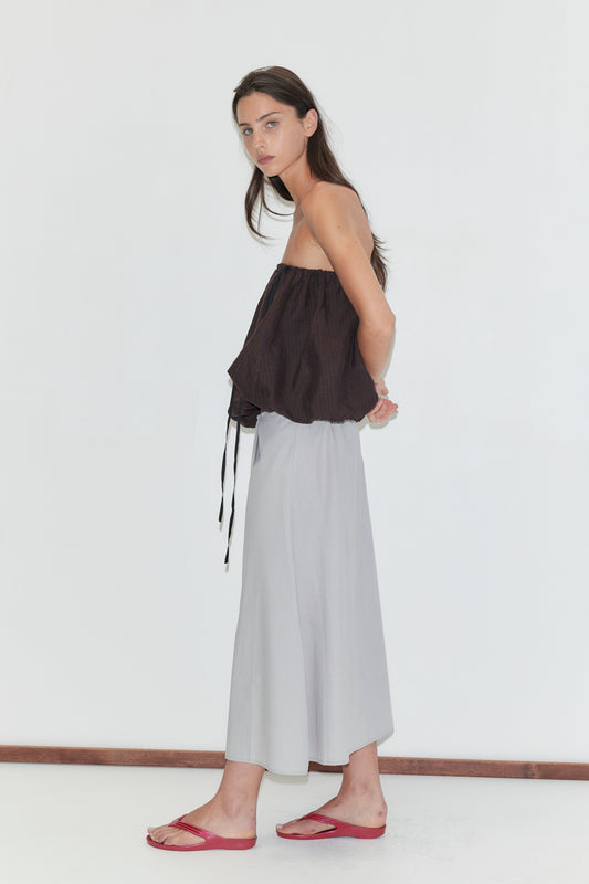 A woman with long dark hair stands in profile against a plain white background wearing the Wrap Skirt - Cloud Stripe by Deiji Studios, a pale grey-blue fine-striped wrap midi skirt, with a brown strapless bubble top and red flip-flops.