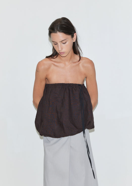 A woman with long dark hair looks down, wearing the Bubble Top - Brown Black Stripe by Deiji Studios, a strapless linen top with an elasticated top edge, contrast black drawcord tie and a bubble hem, paired with a light grey striped wrap skirt against a plain white background.