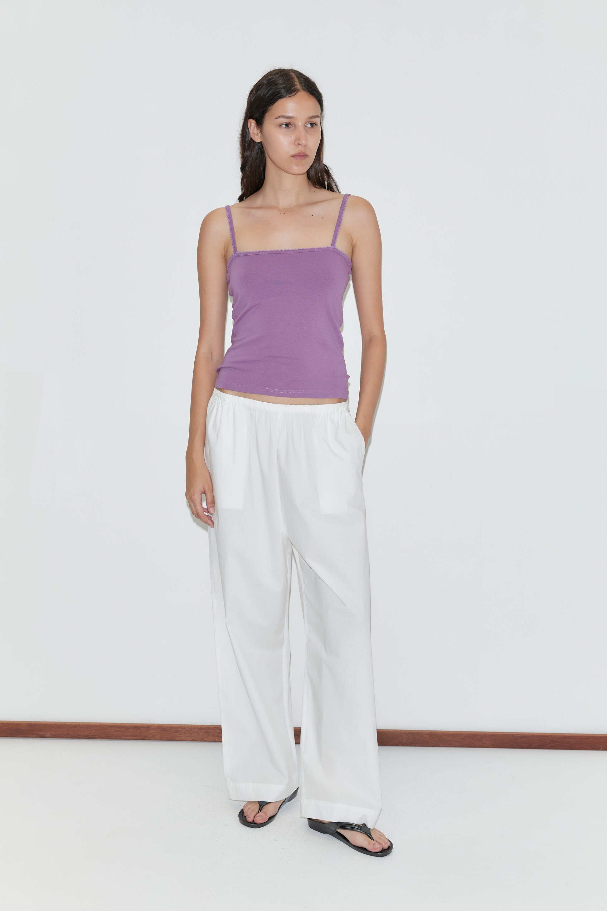 A woman with long dark hair stands against a white background wearing the Scallop Tank - Berry by Deiji Studios, loose white pants, and black sandals. She has one hand in her pocket and looks slightly to the side.