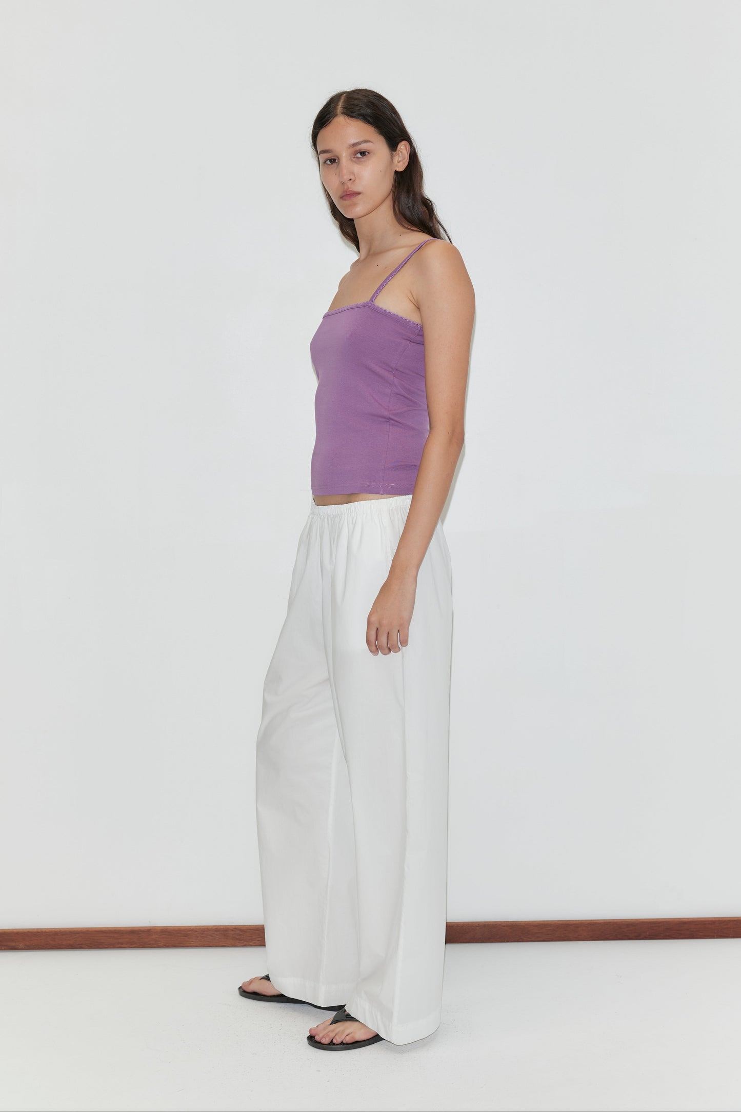 A woman stands against a plain white background, wearing the Scallop Tank - Berry by Deiji Studios, loose white pants, and black sandals. She has long brown hair, a neutral expression, and her arms are relaxed by her sides.
