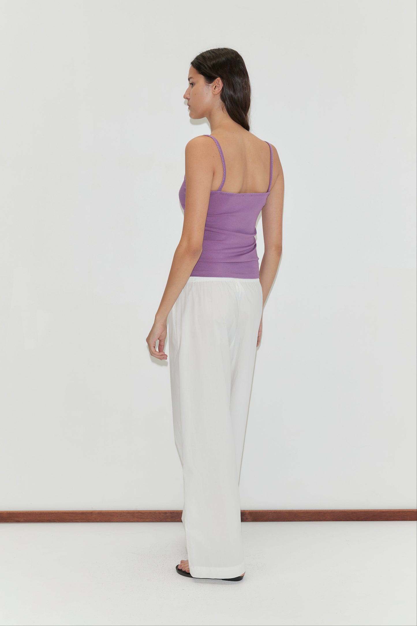 A woman stands sideways to the camera against a plain white background, wearing the Scallop Tank in Berry from Deiji Studios, paired with white loose pants and black sandals.
