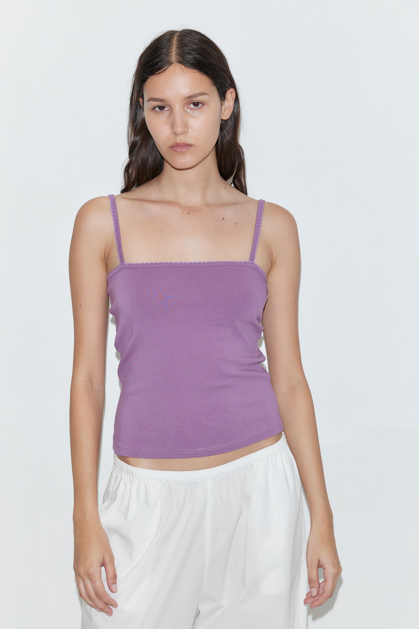 A woman with long dark hair wears a Deiji Studios Scallop Tank in Berry and loose white pants, standing against a plain white background and looking directly at the camera with a neutral expression.
