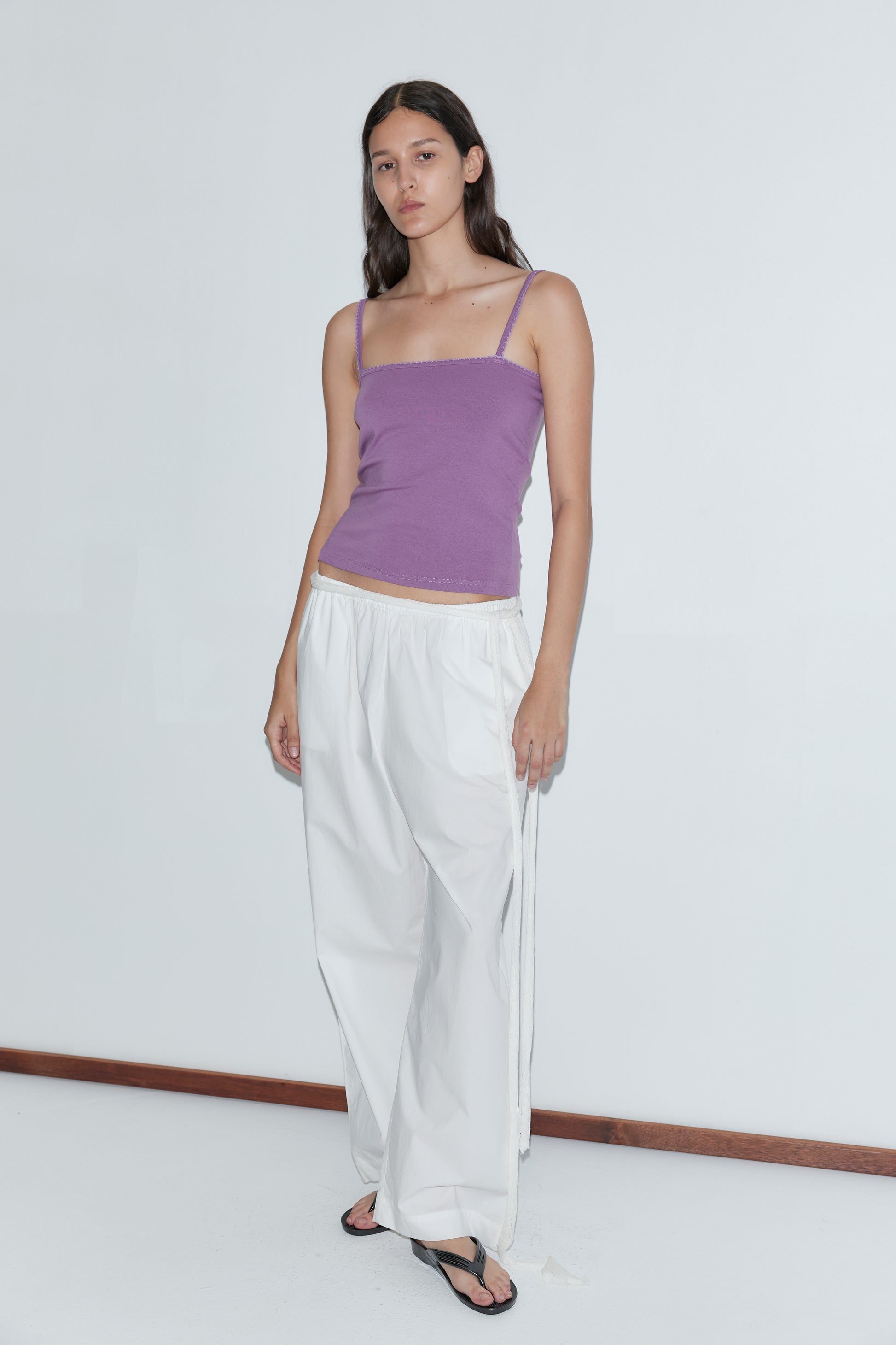 A woman with long brown hair and a neutral expression stands against a plain white wall, wearing the Scallop Tank - Berry, loose white pants from Deiji Studios, and black sandals.