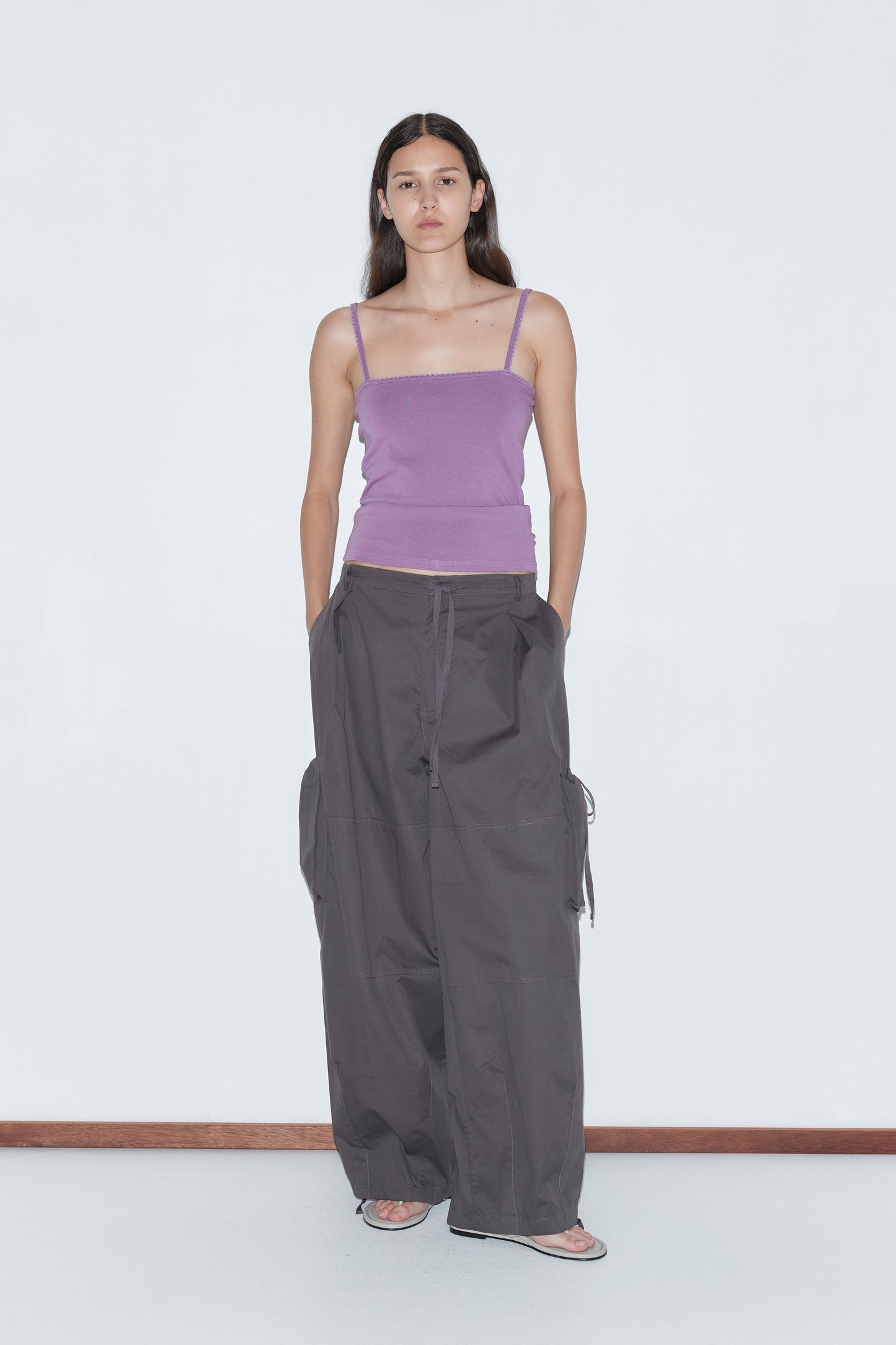 A woman stands against a plain white background, wearing a fitted lavender spaghetti strap top, Deiji Studios Cargo Pant - Carob, and sandals, with her hands in her pockets and a neutral expression.