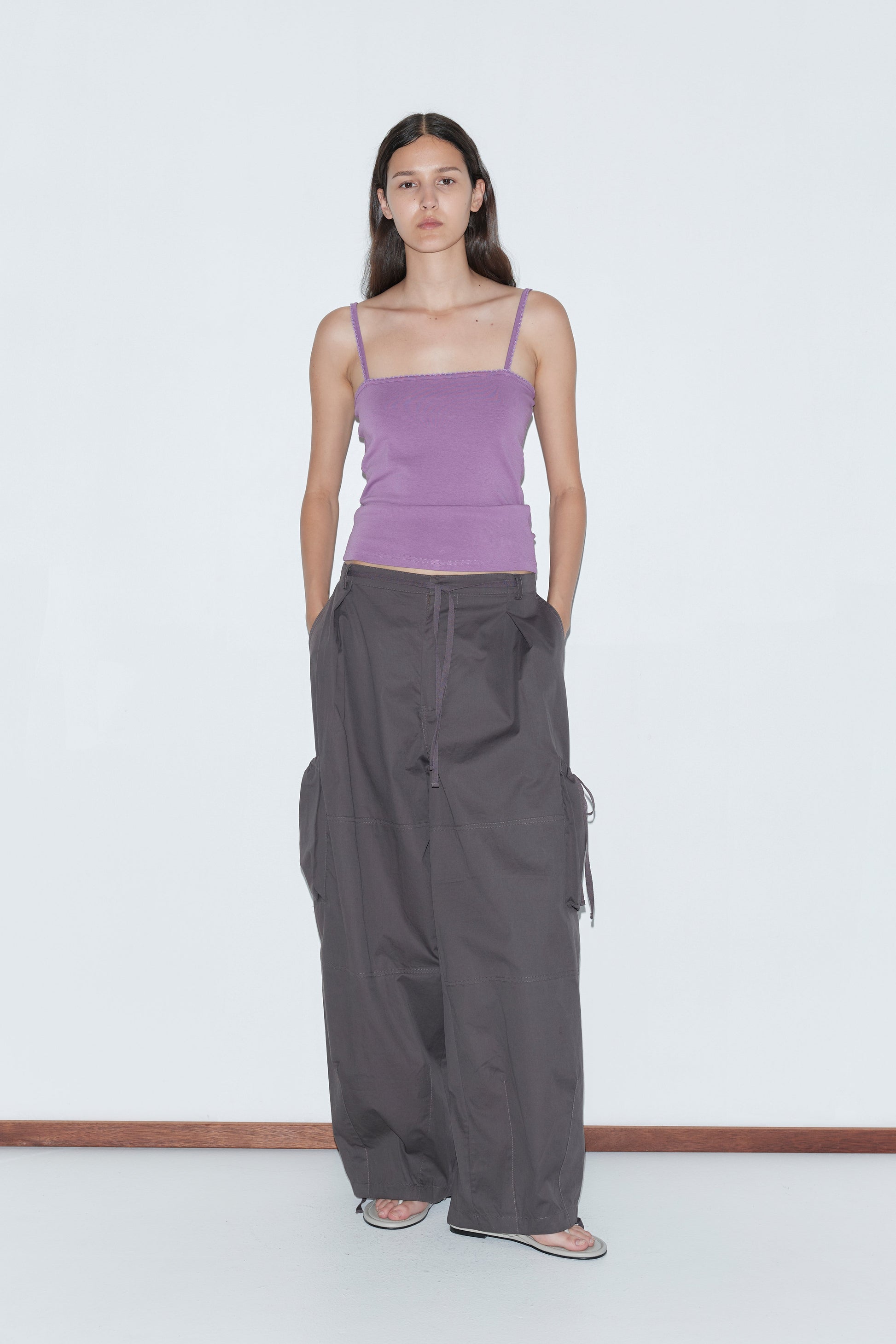 A woman stands against a plain white background, wearing a fitted lavender spaghetti strap top, Deiji Studios Cargo Pant - Carob, and sandals, with her hands in her pockets and a neutral expression.