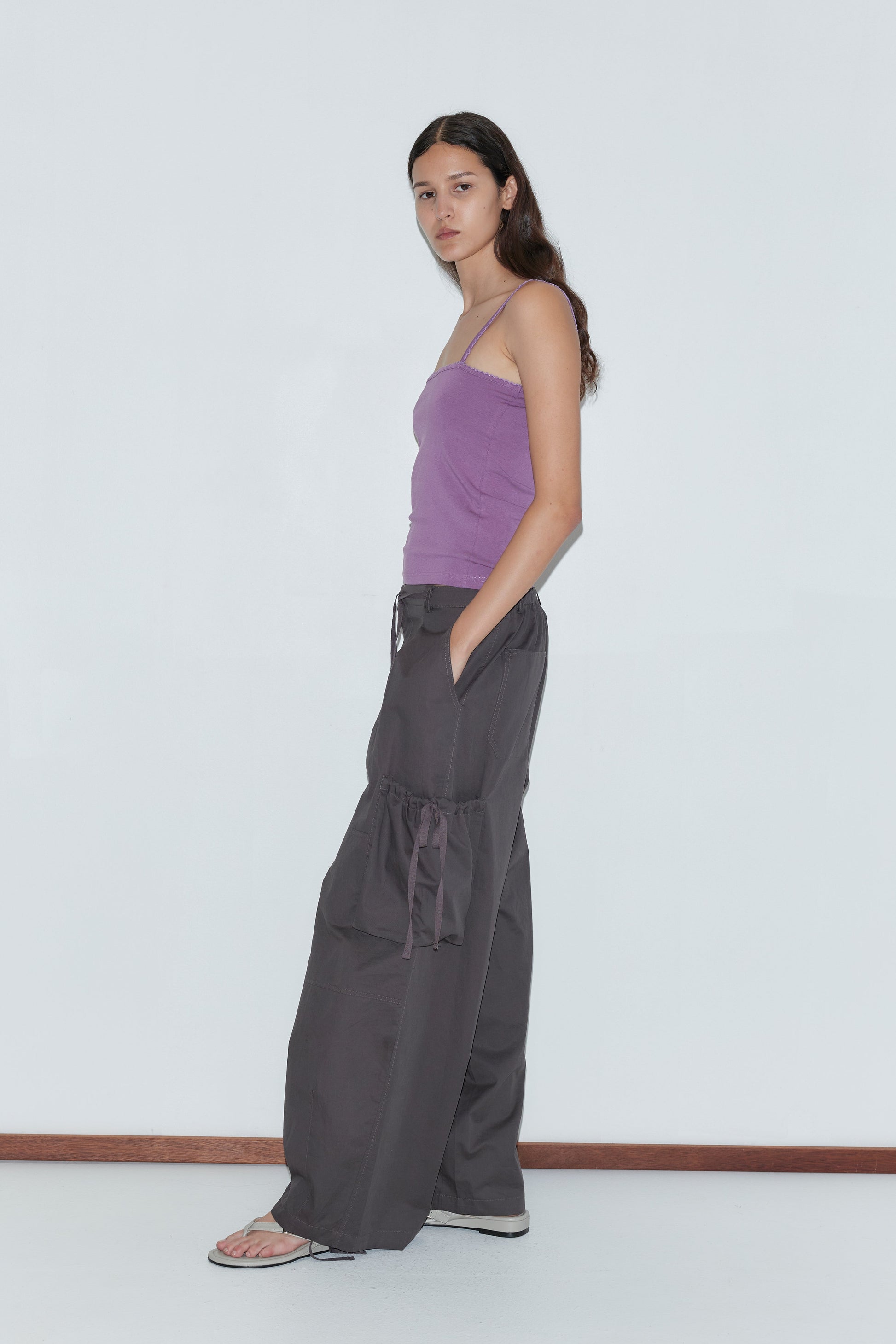 A woman with long dark hair stands against a white wall, wearing a purple spaghetti strap top, the Cargo Pant - Carob by Deiji Studios in loose dark gray with side ties, and white flip-flops. She looks at the camera with a neutral expression.