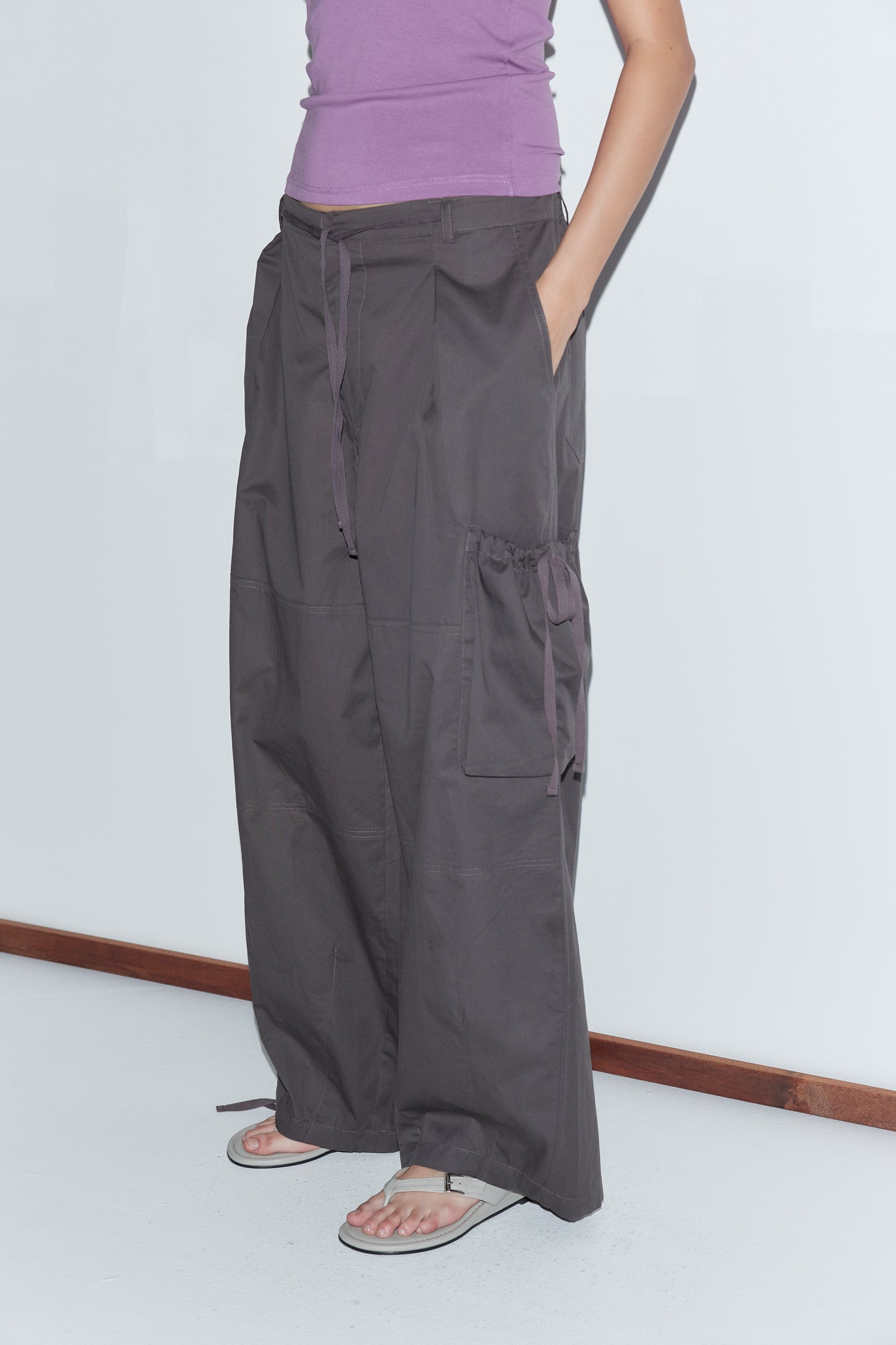 A person wearing a purple tank top, loose deiji studios Cargo Pant - Carob with large pockets, and beige sandals stands against a plain white wall with their hands in their pockets.
