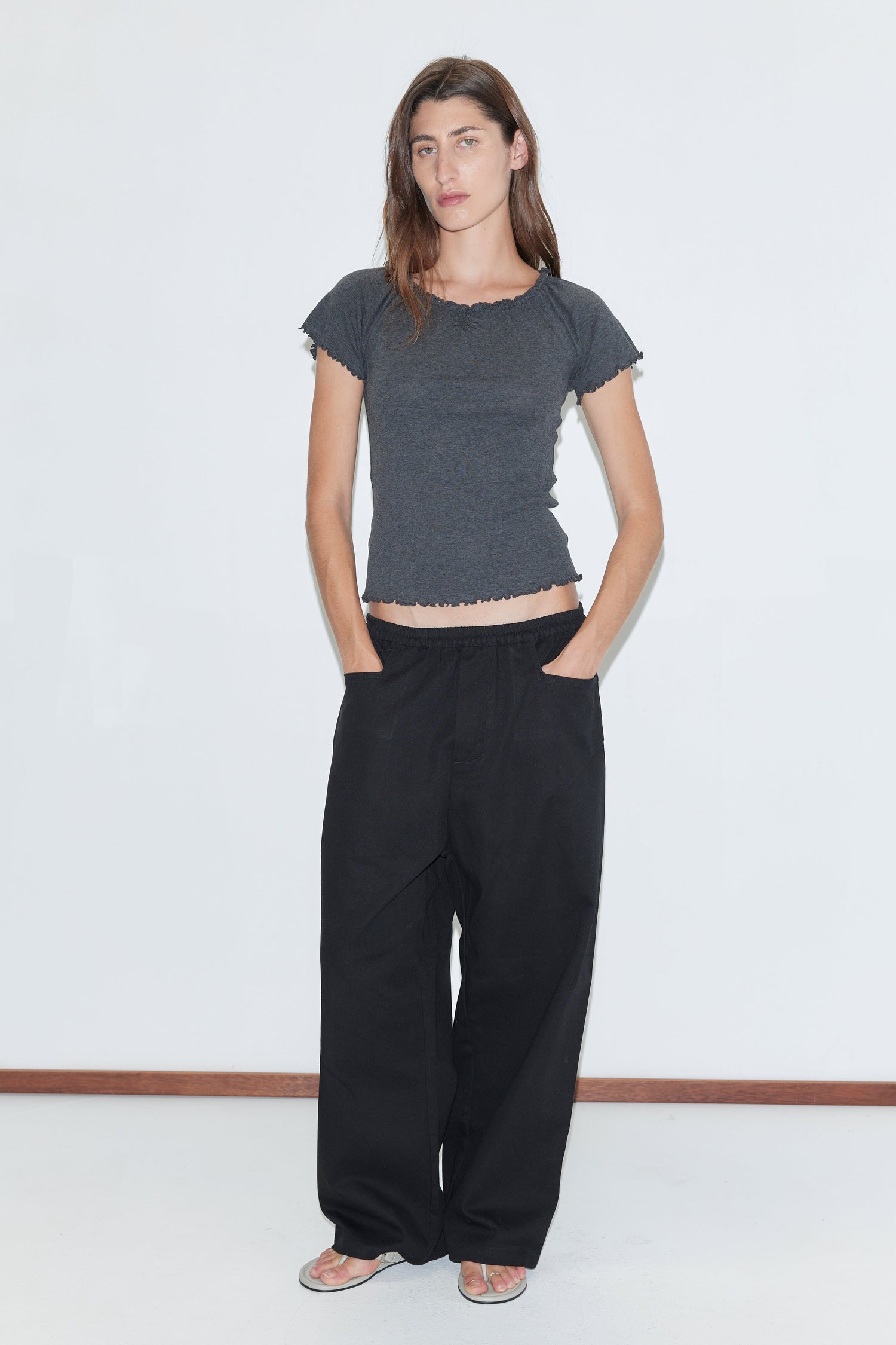 A woman stands against a white wall, wearing a fitted dark gray short-sleeve top, the Square Pocket Pant - Black by Deiji Studios, and sandals. She has long brown hair and her hands are in her pockets.