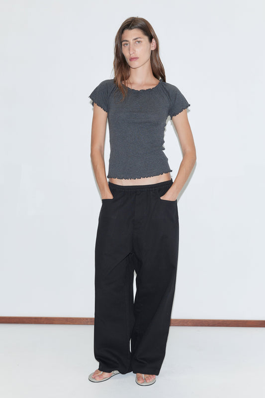 A woman with long brown hair and a neutral expression stands against a white background, wearing the Square Pocket Pant in black—relaxed organic cotton twill pants with square pockets—paired with a fitted dark gray short-sleeved top and flat sandals.