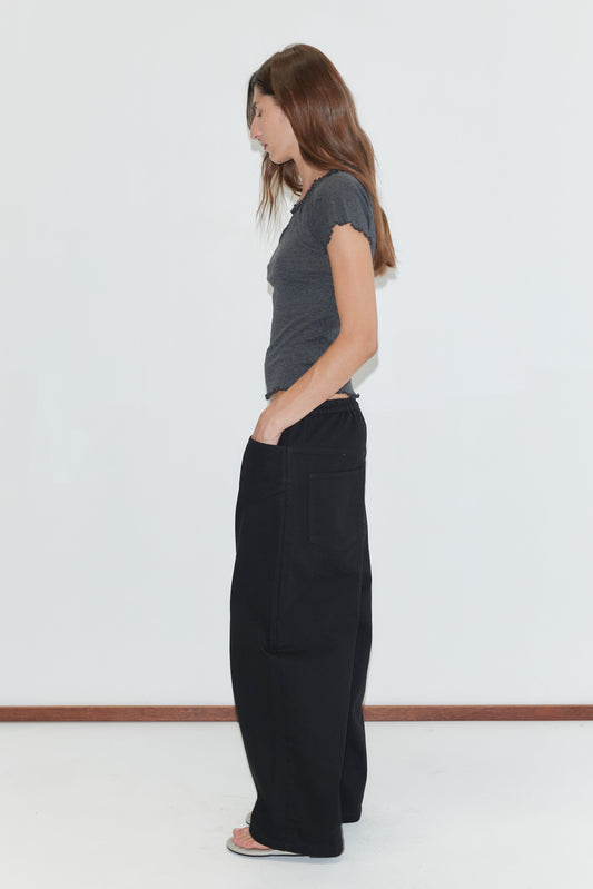 A woman with long brown hair stands sideways against a white background, wearing a fitted gray t-shirt, the Square Pocket Pant - Black in organic cotton twill, and sandals.