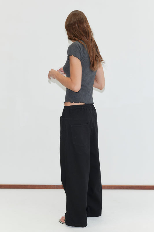 A woman with long brown hair, wearing a fitted gray top and the Square Pocket Pant - Black, stands facing away from the camera against a plain white background.