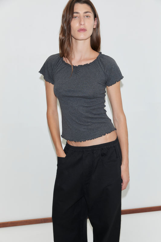 A woman with long brown hair wears a fitted, short-sleeve dark gray top and the Square Pocket Pant in Black, her hands in the pockets as she stands against a plain white background.