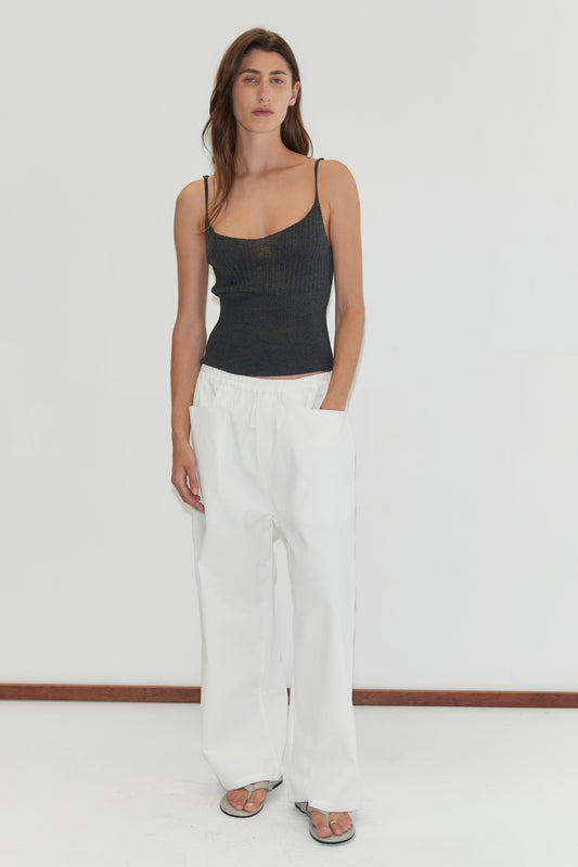 A woman with long brown hair wears a black spaghetti strap top, Square Pocket Pant in white, and sandals, standing against a plain white background.