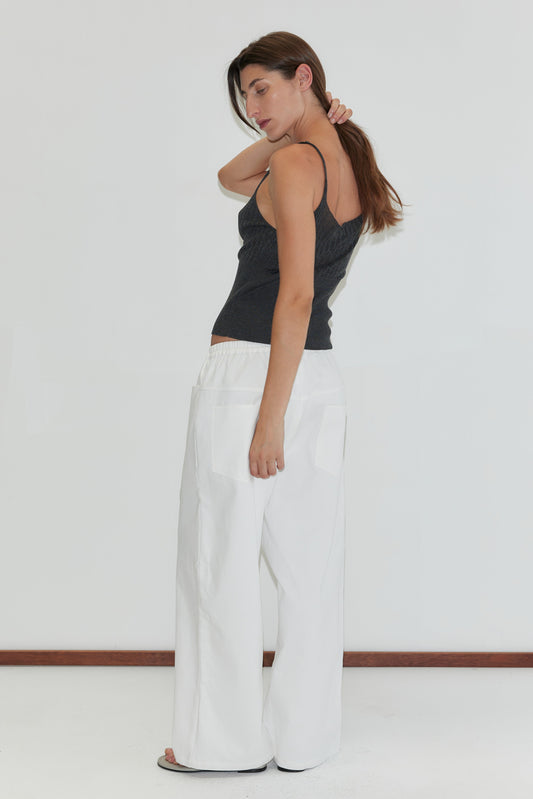 A woman stands sideways against a plain white background, wearing the Square Pocket Pant in White. She has long brown hair, is looking down, and touches the back of her neck with one hand.