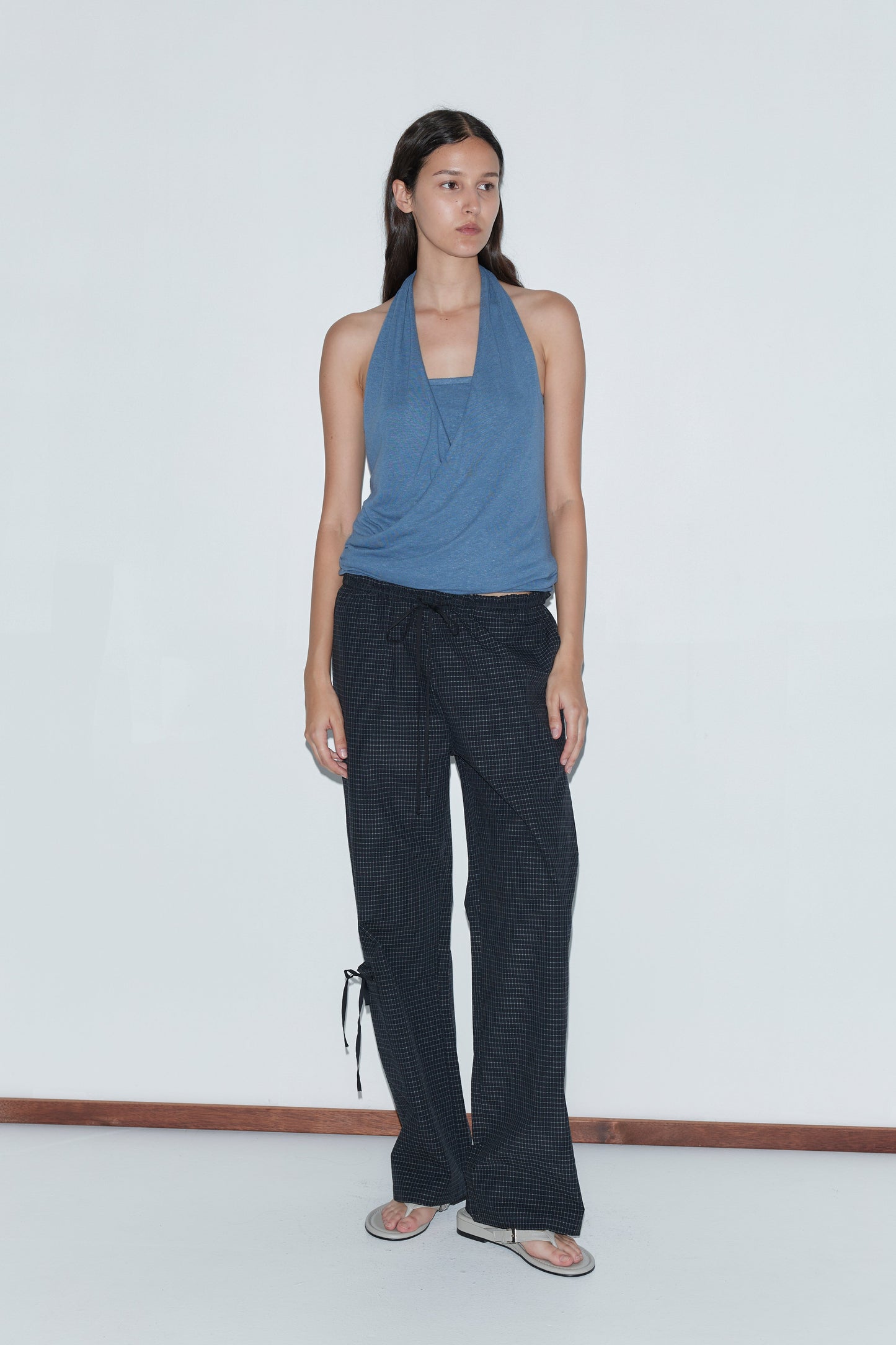 A woman with long dark hair stands against a white wall wearing the Double Layer Jersey Halter - Blue by Deiji Studios, loose black patterned pants, and flip-flops, gazing slightly to her left with a neutral expression.