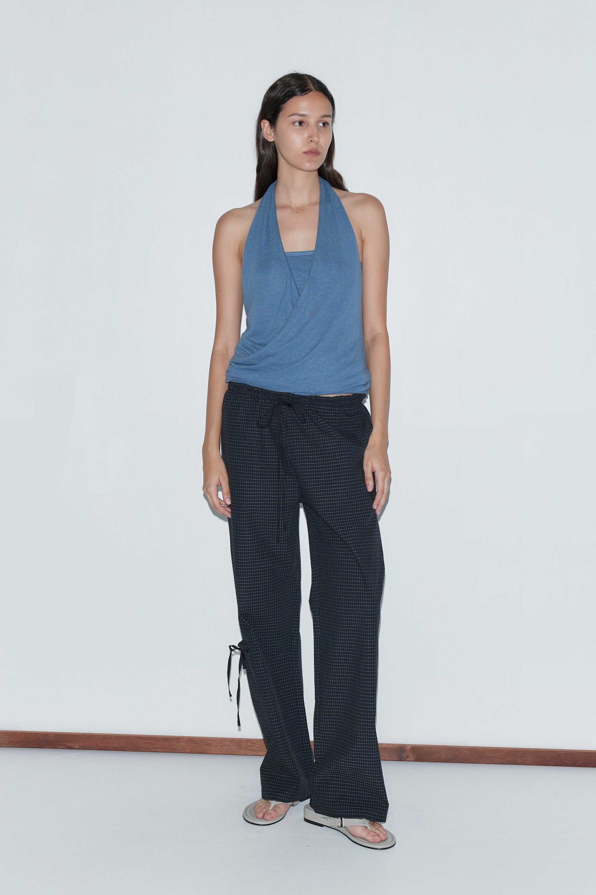 A woman with long dark hair stands against a white wall wearing the Double Layer Jersey Halter - Blue by Deiji Studios, loose black patterned pants, and flip-flops, gazing slightly to her left with a neutral expression.