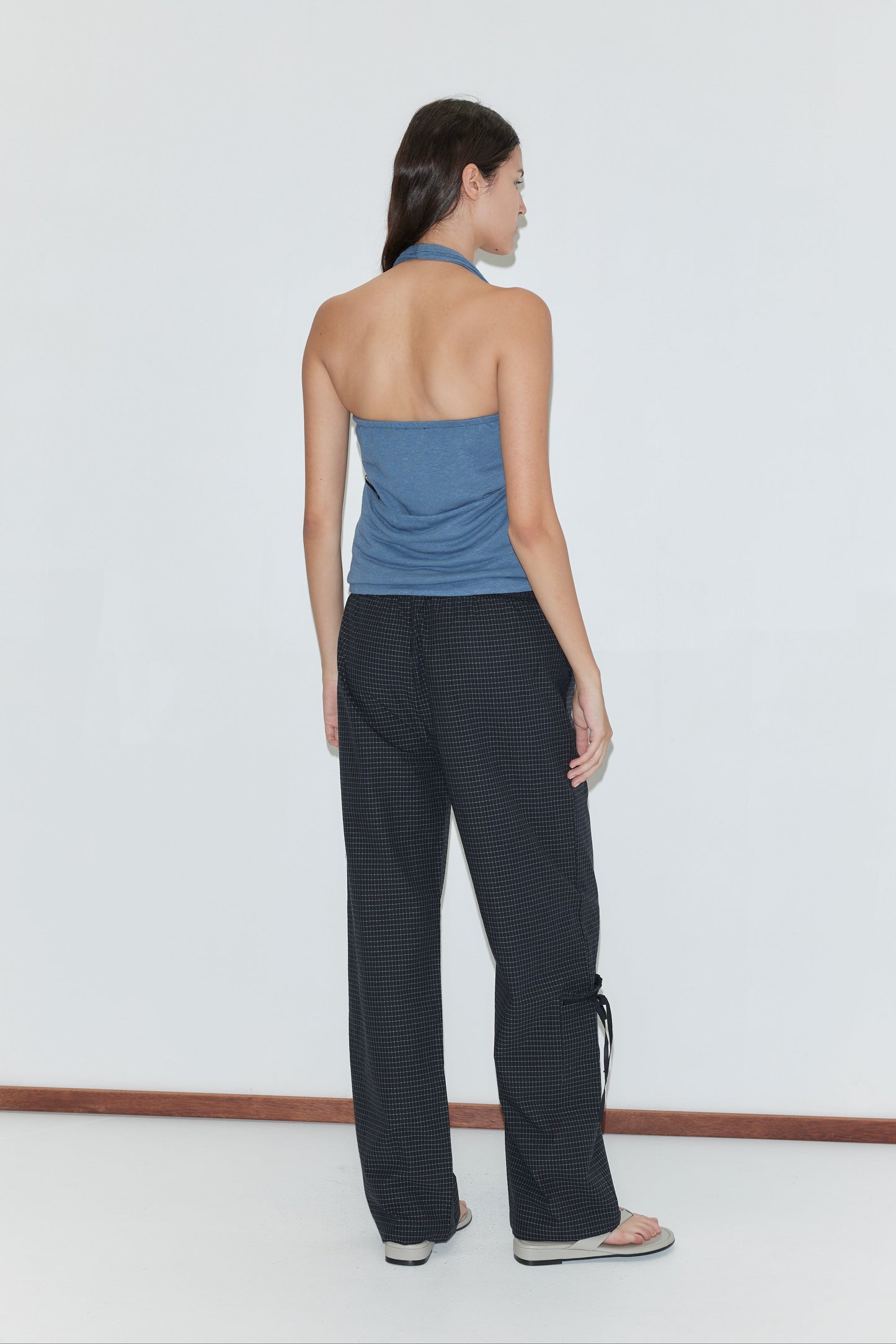 A woman faces away, wearing a blue halter top, Grosgrain Pant - Mono Grid by Deiji Studios, and flip-flops against a plain white background.
