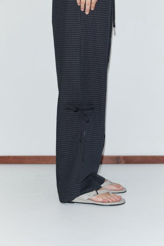A person stands on a white floor wearing Grosgrain Pant - Mono Grid and white flip-flops. Only the lower half of their body is visible, with one hand resting by their side.