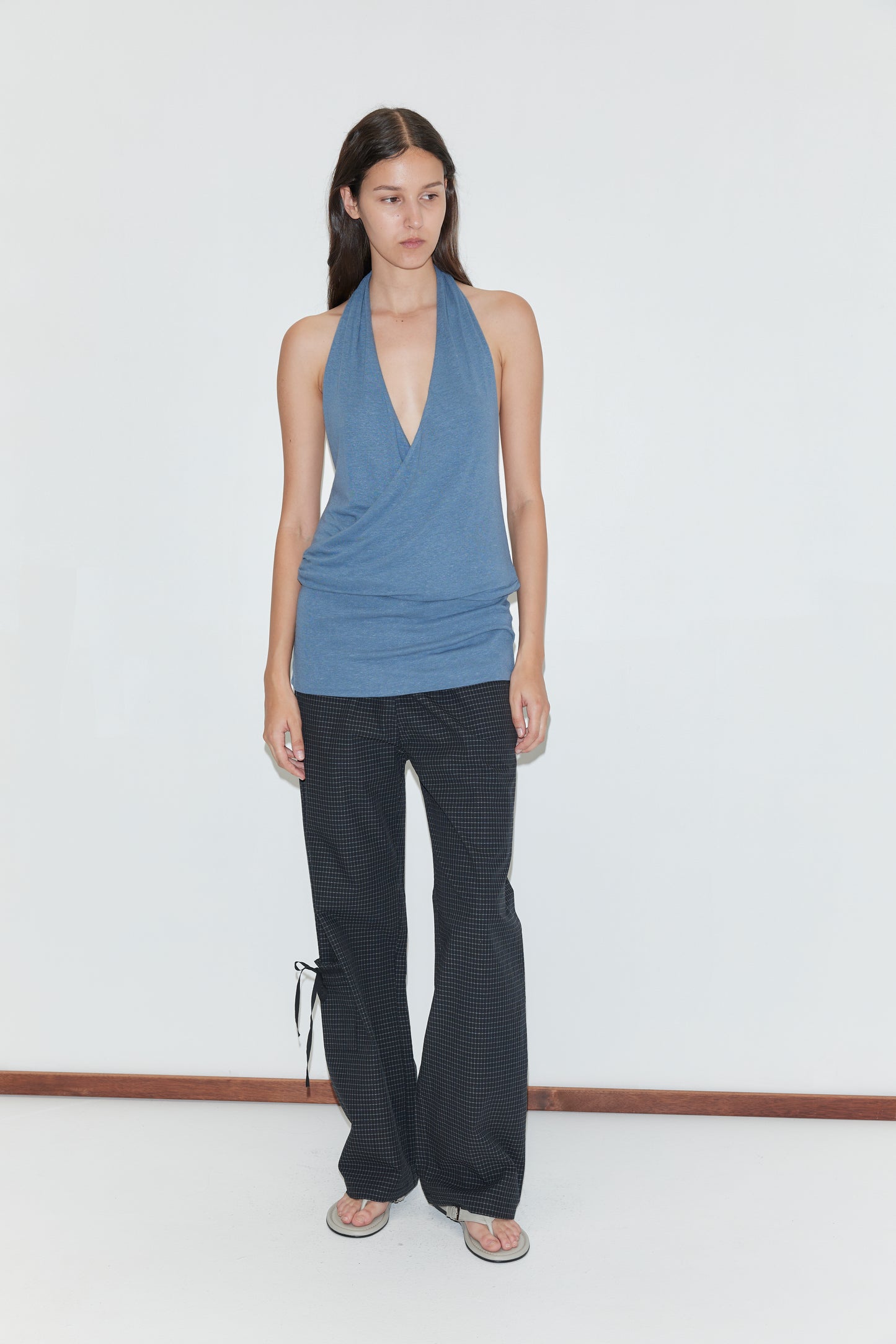 A person with long dark hair and a neutral expression stands against a white wall, wearing the Double Layer Jersey Halter - Blue, black patterned flared pants with side ties, and flip-flops.