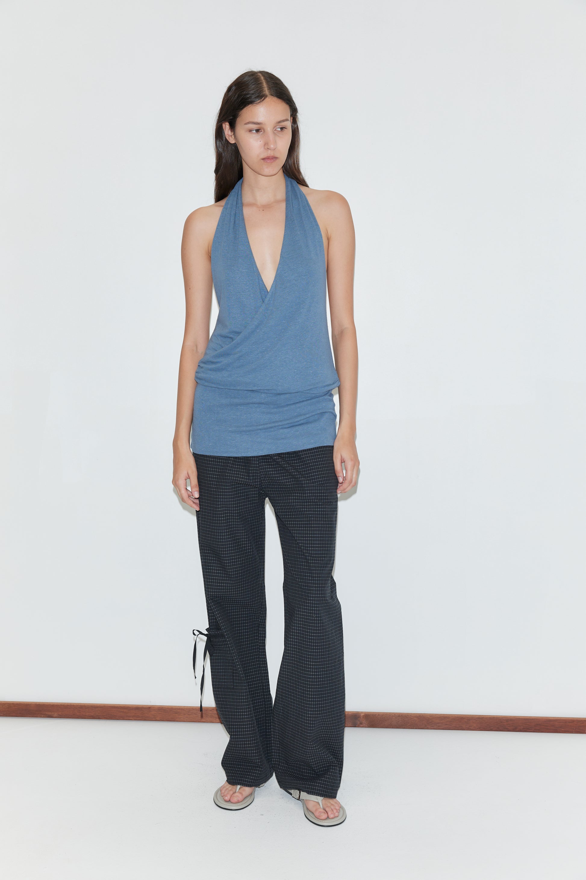 A person with long dark hair and a neutral expression stands against a white wall, wearing the Double Layer Jersey Halter - Blue, black patterned flared pants with side ties, and flip-flops.