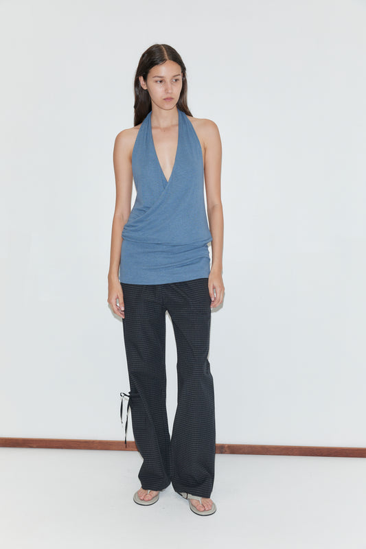 A person with long dark hair and a neutral expression stands against a white wall, wearing the Double Layer Jersey Halter - Blue, black patterned flared pants with side ties, and flip-flops.