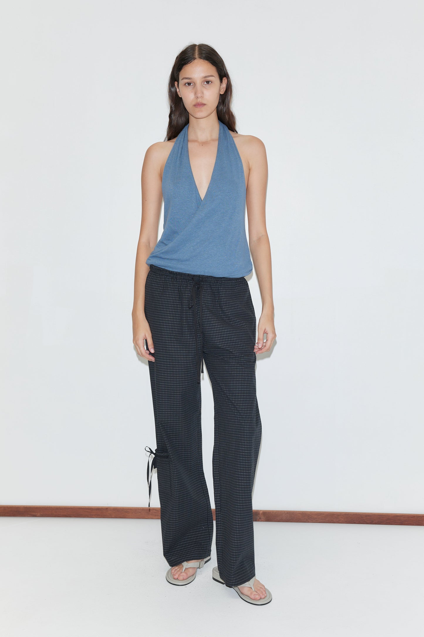 A person with long dark hair and a neutral expression stands against a white wall wearing a blue halter top, flip-flops, and Deiji Studios’ Grosgrain Pant - Mono Grid featuring relaxed legs and ankle ties.