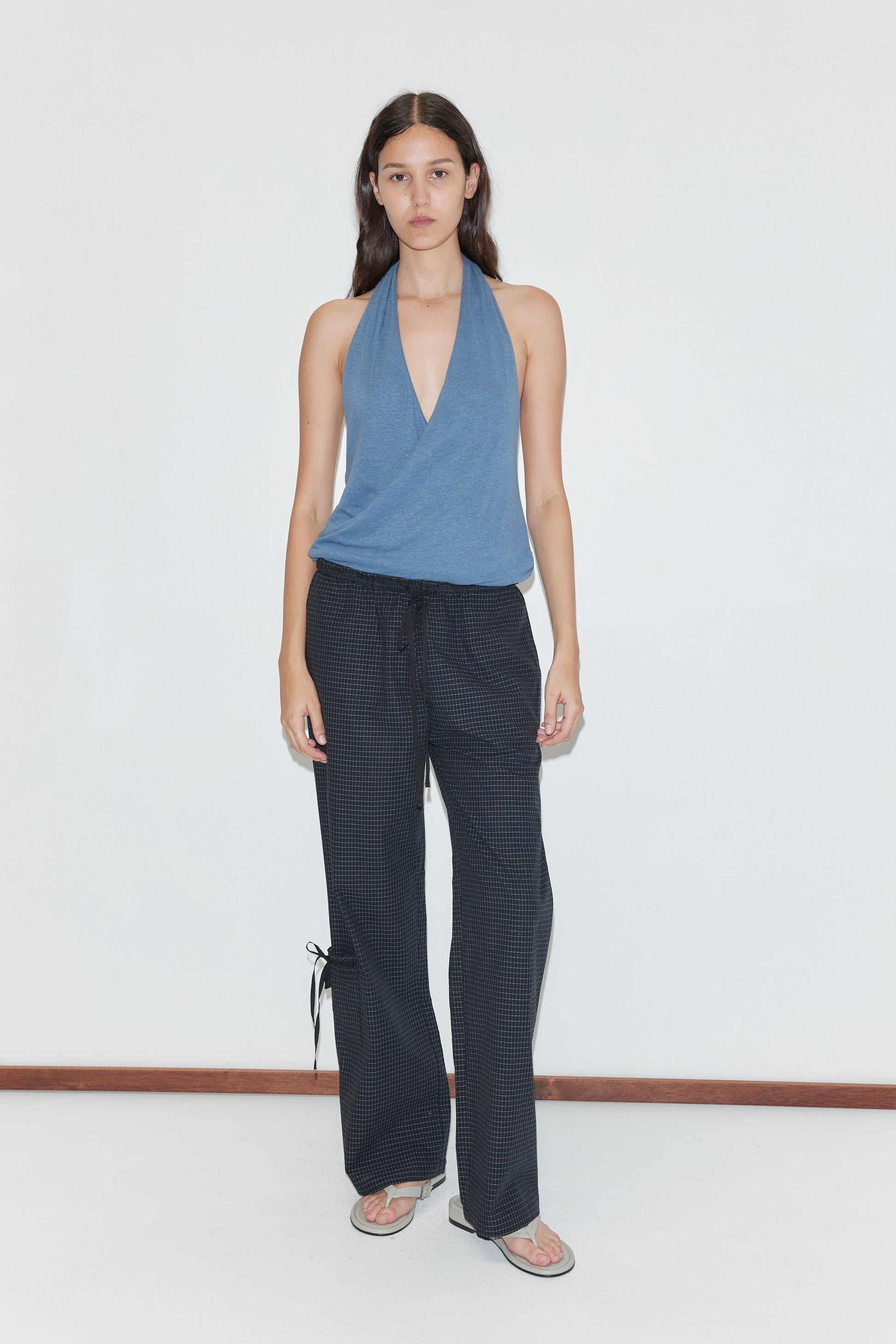 A person with long dark hair and a neutral expression stands against a white wall wearing a blue halter top, flip-flops, and Deiji Studios’ Grosgrain Pant - Mono Grid featuring relaxed legs and ankle ties.