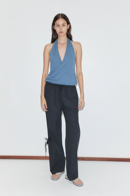 A person with long dark hair and a neutral expression stands against a white wall wearing a blue halter top, flip-flops, and Deiji Studios’ Grosgrain Pant - Mono Grid featuring relaxed legs and ankle ties.
