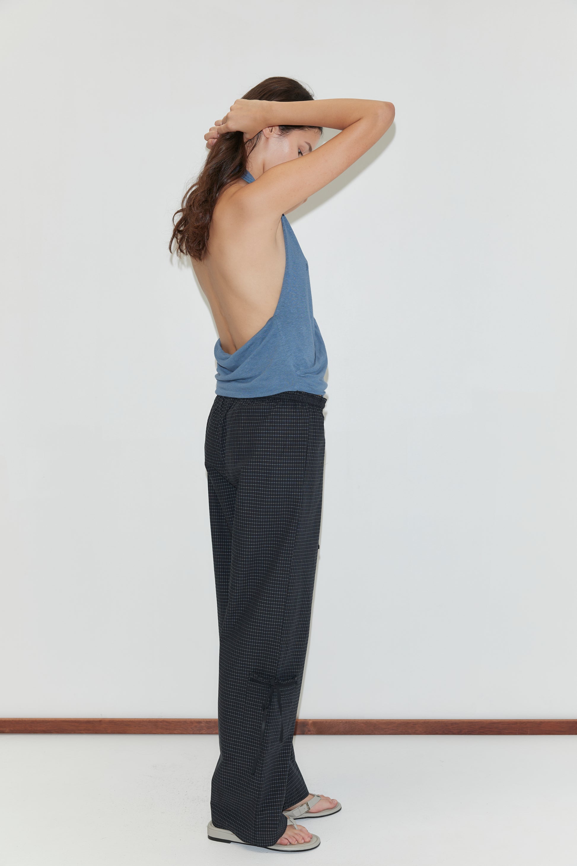A person stands in profile against a white background, wearing a loose blue sleeveless top with an open back, Grosgrain Pant - Mono Grid, and sandals, arms raised and hands touching the back of their head.