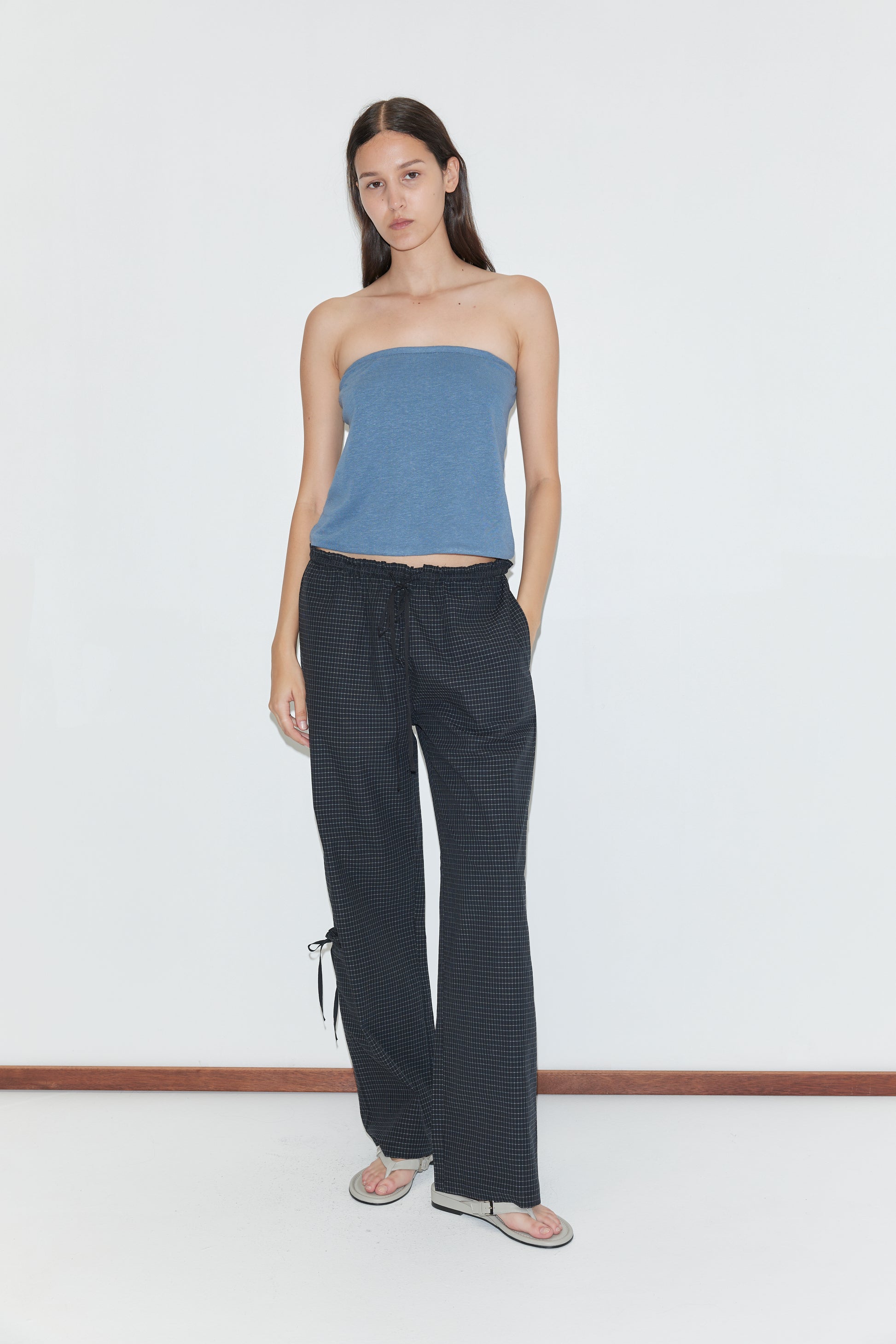 A woman stands against a plain white wall, wearing a blue strapless top, Grosgrain Pant - Mono Grid by Deiji Studios with a subtle pattern, and flip-flops. Her hands are in her pockets and she has long, straight hair.