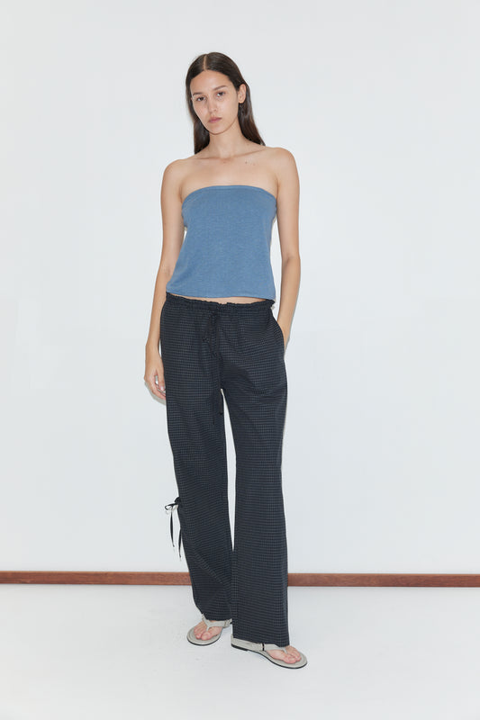 A woman stands against a plain white wall, wearing a blue strapless top, Grosgrain Pant - Mono Grid by Deiji Studios with a subtle pattern, and flip-flops. Her hands are in her pockets and she has long, straight hair.