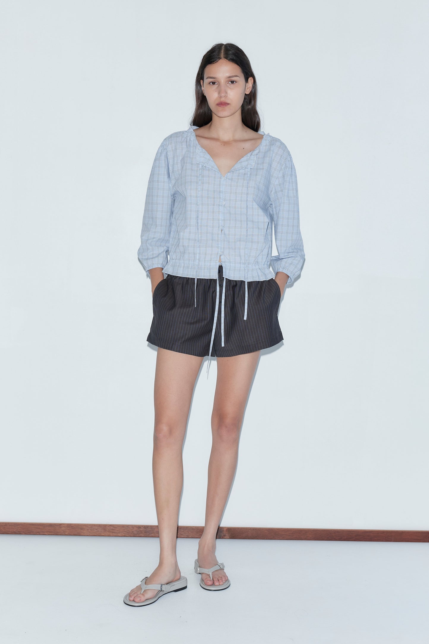 A woman with long dark hair stands against a white background, wearing the Corded Top - Powder Check by Deiji Studios, paired with black shorts with white drawstrings and flip-flops. Her hands are in her pockets.