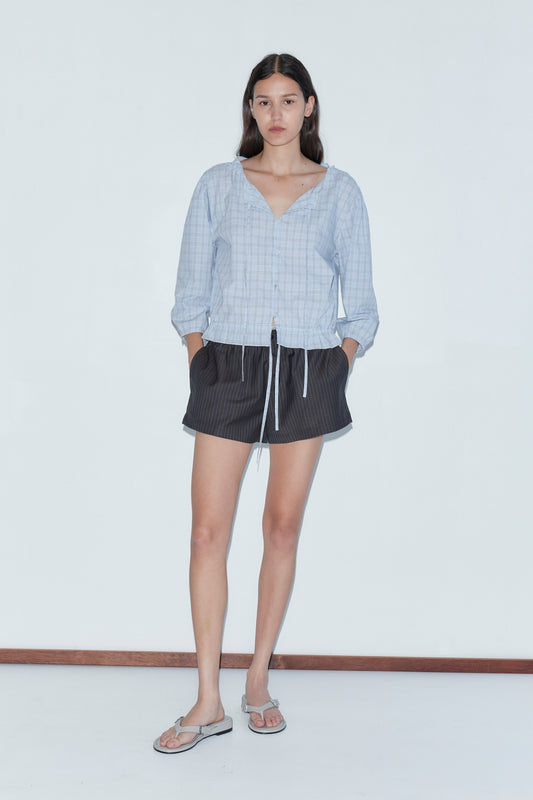 A woman with long dark hair stands against a white background, wearing the Corded Top - Powder Check by Deiji Studios, paired with black shorts with white drawstrings and flip-flops. Her hands are in her pockets.