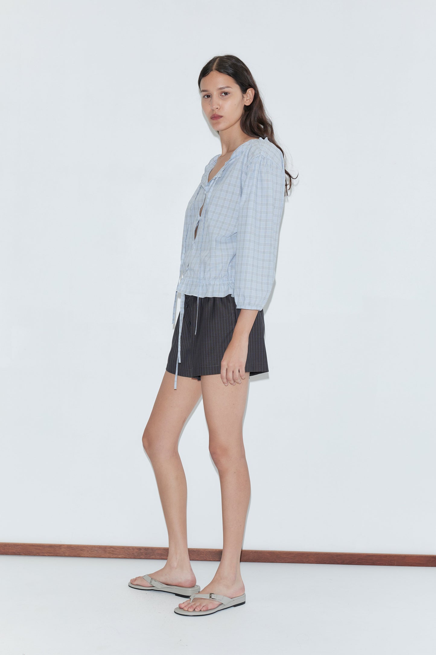 A young woman with long, dark hair stands in profile against a white background, wearing the Corded Top - Powder Check by deiji studios, paired with dark shorts and flip-flops. She maintains a neutral expression.